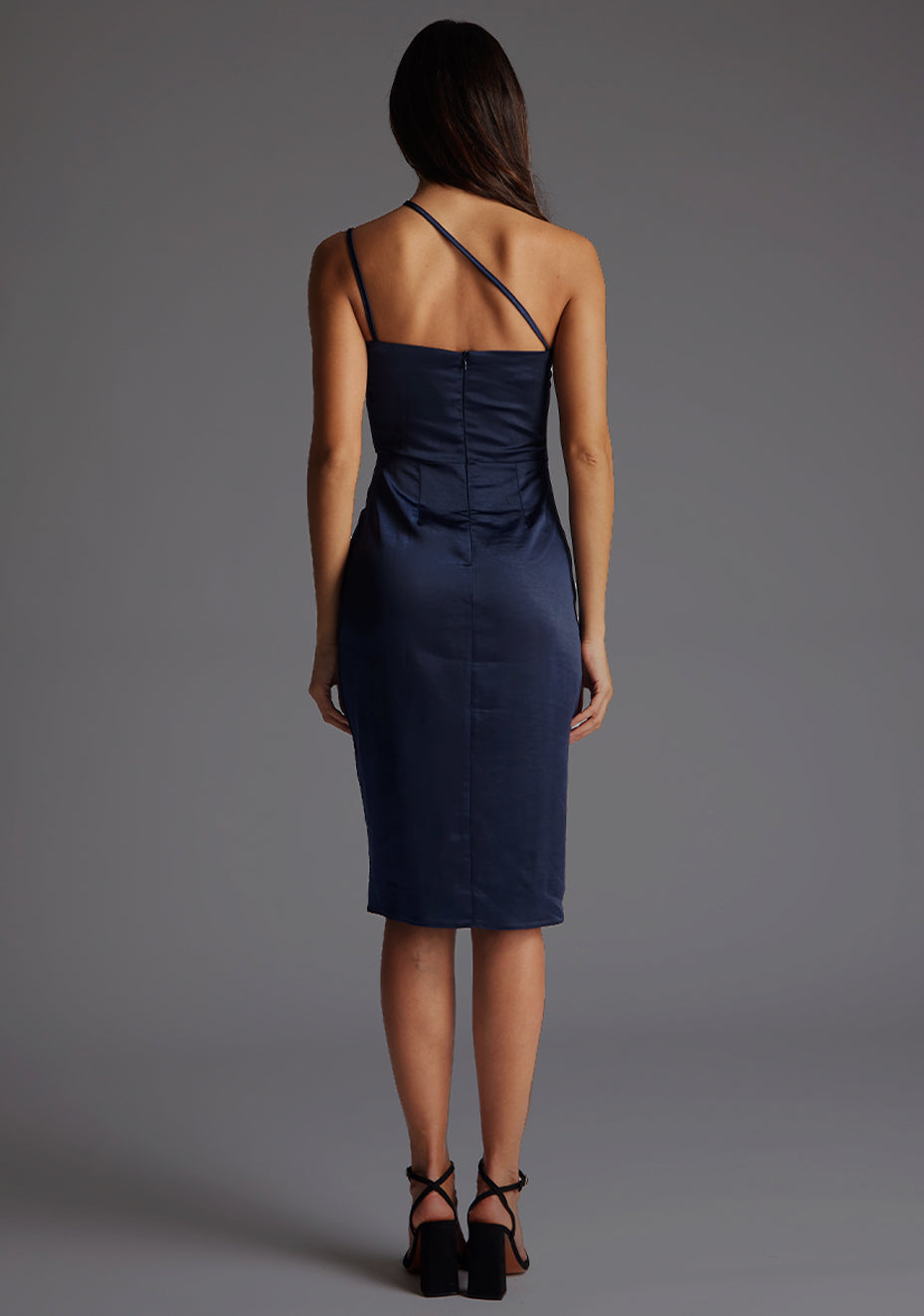 Back image of a model wearing a navy satin midi dress with a straight neckline with a asymmetric strap across front of chest and invisible centre back zip. The design featured is the Vesper Rosalina Navy Satin Midi Dress