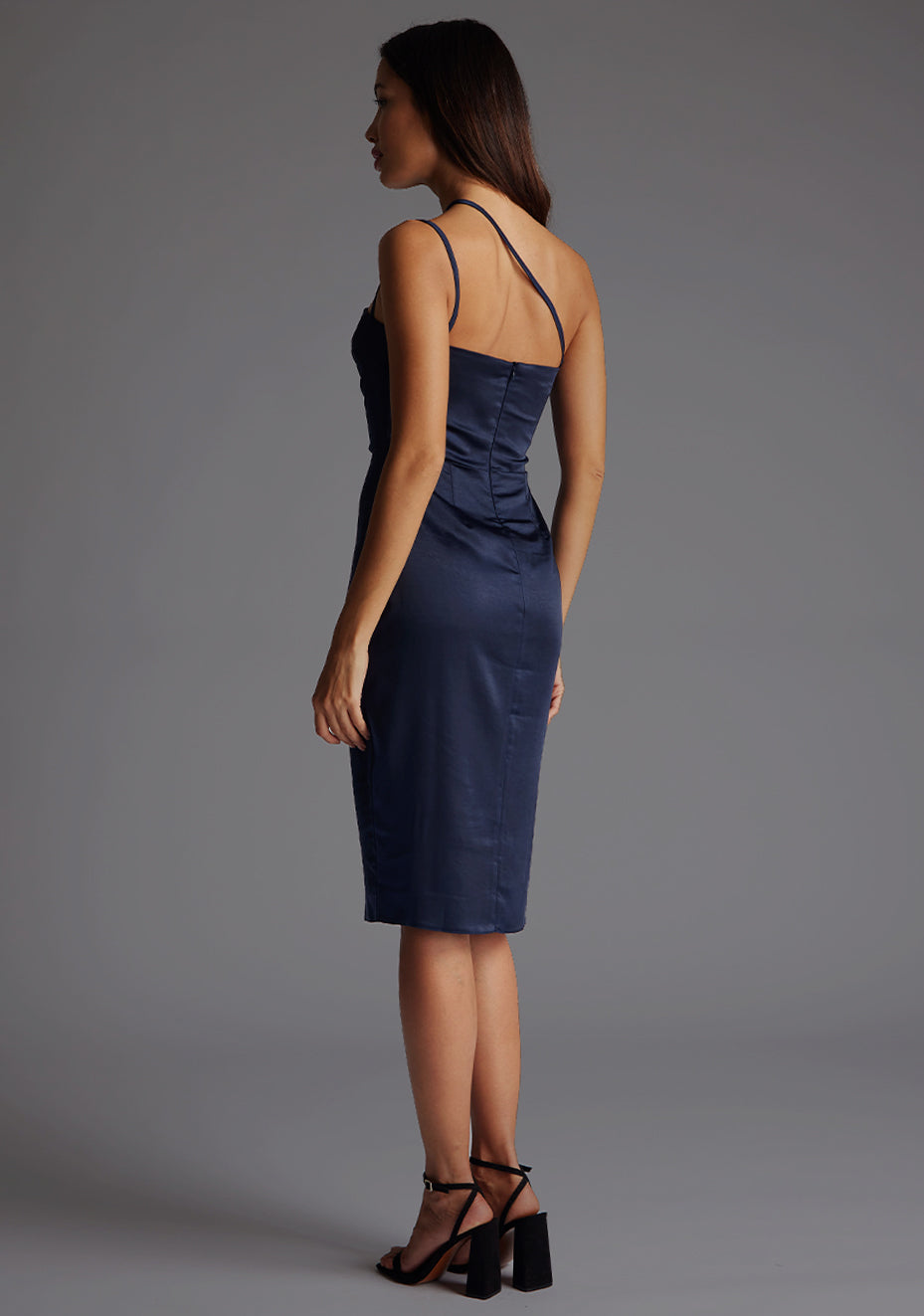 Back quarter image of a model wearing a navy satin midi dress with a straight neckline with a asymmetric strap across front of chest and invisible centre back zip. The design featured is the Vesper Rosalina Navy Satin Midi Dress