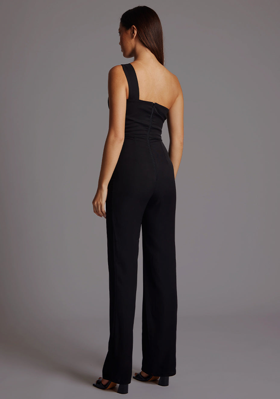 Back image of a model wearing a black Jumpsuit with a stylish one-shoulder design, accentuated by a sophisticated front cut-out, slim fit and convenient back zip. The design featured is the Vesper Rosemary Black Jumpsuit.