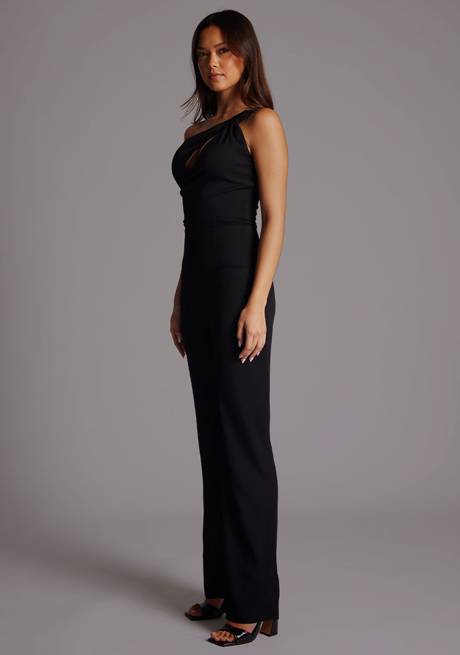 Front quarter image of a model wearing a black Jumpsuit with a stylish one-shoulder design, accentuated by a sophisticated front cut-out, slim fit and convenient back zip. The design featured is the Vesper Rosemary Black Jumpsuit.
