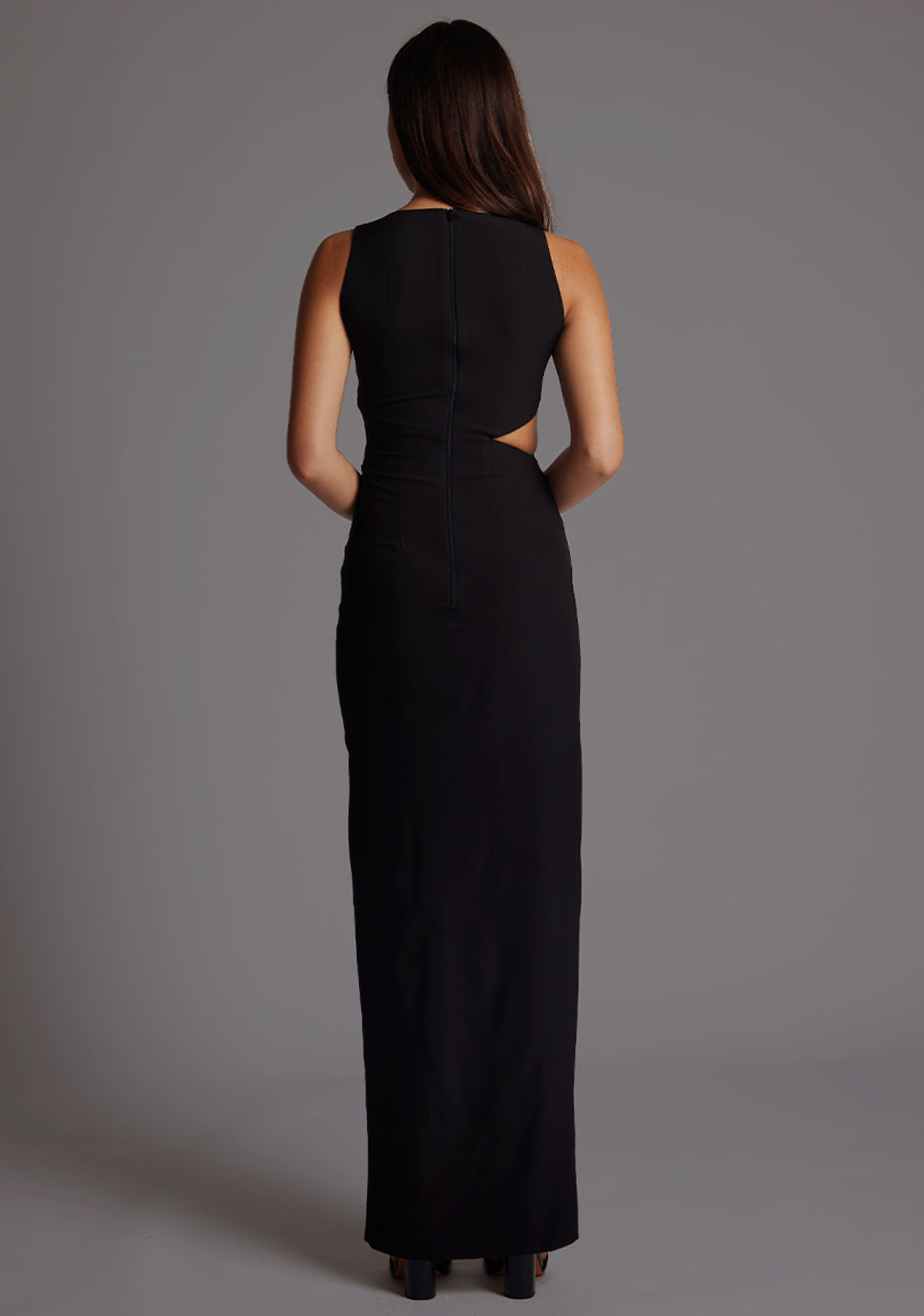 Back image of a model wearing a black maxi dress, featuring a round neckline, side bodice cut-out, side waist pleating detail and front skirt split. The dress featured is the Vesper Rowan Black Maxi Cut Out Dress.