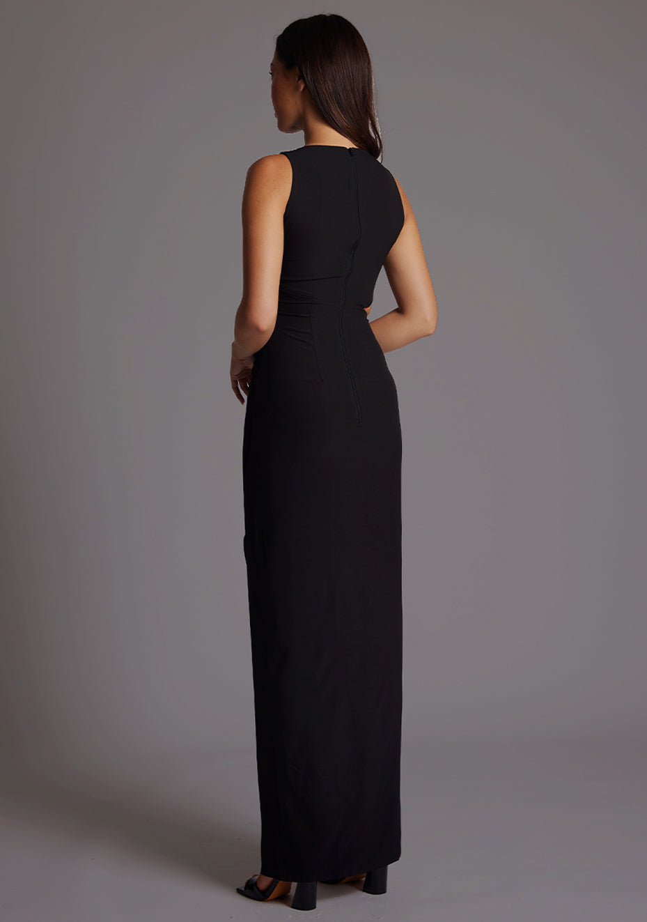 Back quarter image of a model wearing a black maxi dress, featuring a round neckline, side bodice cut-out, side waist pleating detail and front skirt split. The dress featured is the Vesper Rowan Black Maxi Cut Out Dress.