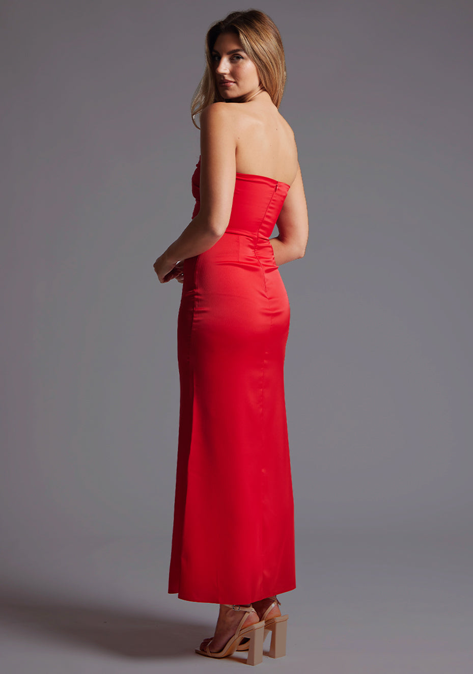 Back quarter image of a blonde model wearing a red midaxi dress with a strapless design, front cutout with a sleek and slim fit silhouette. The design featured is the Vesper Sabine Red Midaxi Dress.