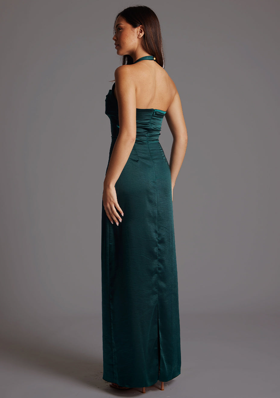 Back quarter image of a model wearing a pine maxi dress with a halter cowl neck exudes sophistication, while the lustrous satin fabric offers a luxurious touch. The design featured is the Vesper Saige Pine Satin Green Maxi Dress.