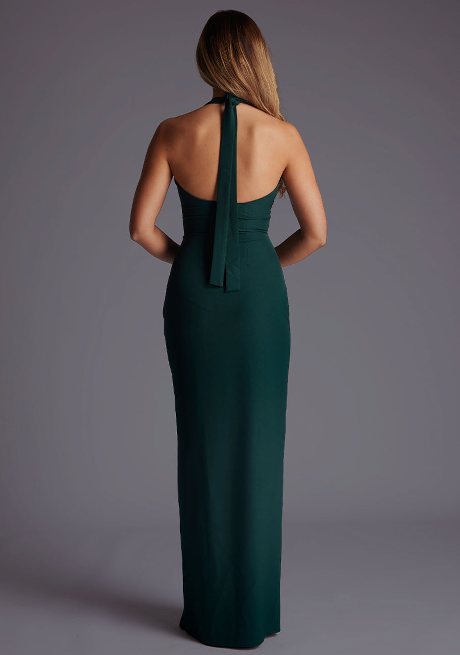 Back image of a blonde model wearing a forest green maxi dress with a v-plunging neckline, halter design and a front skirt split. The design featured is the Vesper Sedona Forest Green Maxi Dress.