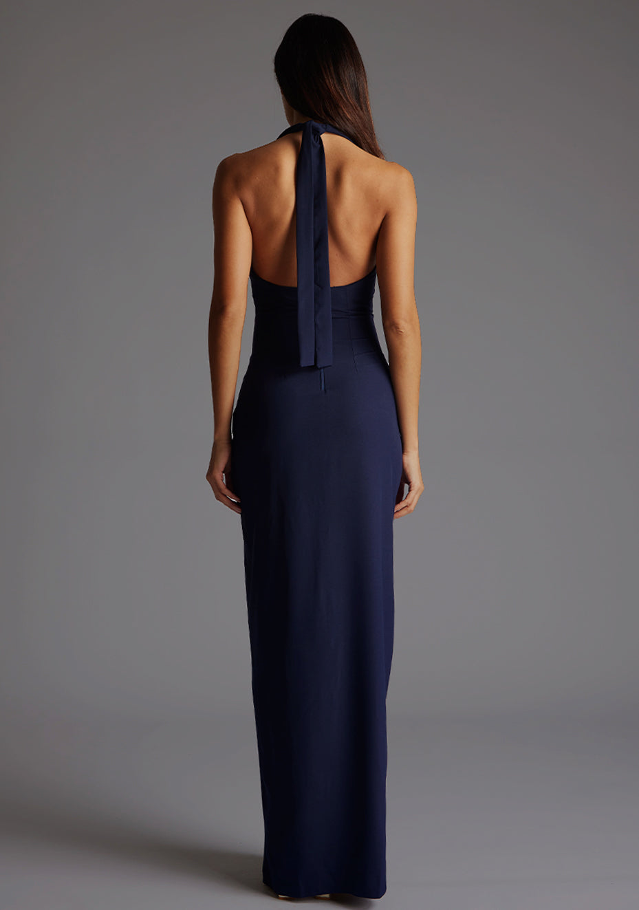 Back image of a blonde model wearing a navy maxidress with a v-plunging neckline, halter design and a front skirt split. The design featured is the Vesper Sedona Navy Maxi Dress.
