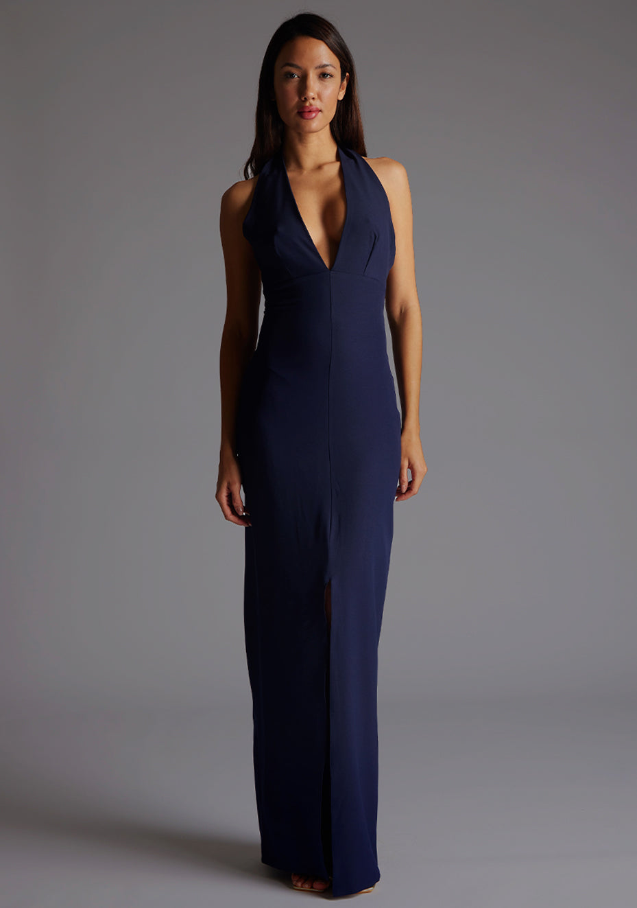 Front image of a blonde model wearing a navy maxidress with a v-plunging neckline, halter design and a front skirt split. The design featured is the Vesper Sedona Navy Maxi Dress