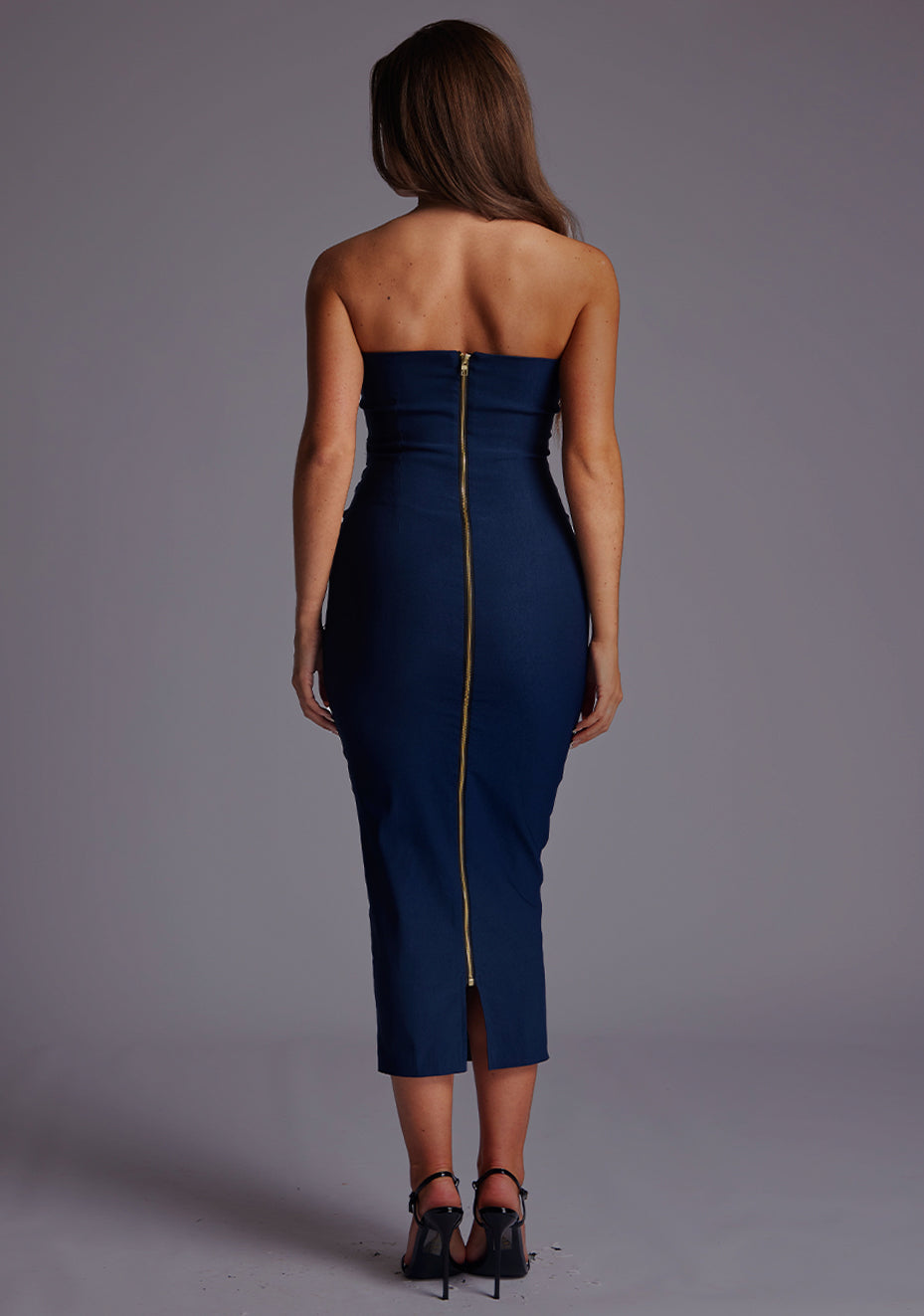 Back image of a model wearing a navy midaxi dress, featuring a strapless neckline with gold zip at the back. The design featured is the Vesper Shelbie Navy Strapless Midaxi Dress.