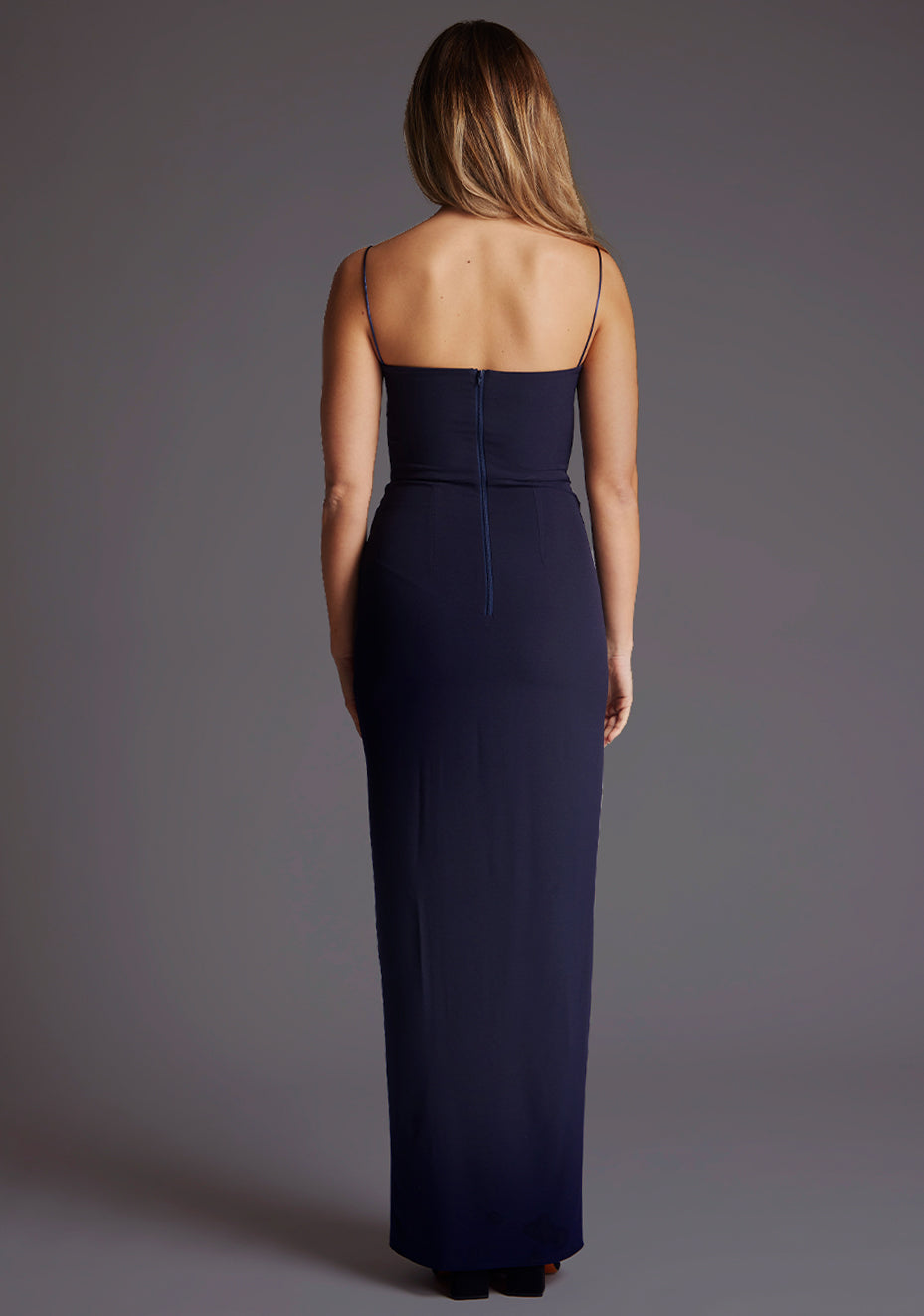 Back image of a model wearing a navy maxi dress, featuring delicate straps and a front skirt split. The design featured is the Vesper Tate Navy Maxi Dress.