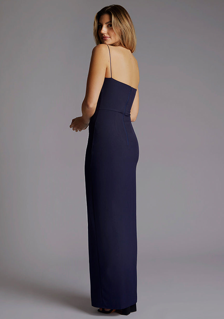 Back quarter image of a model wearing a navy maxi dress, featuring delicate straps and a front skirt split. The design featured is the Vesper Tate Navy Maxi Dress.