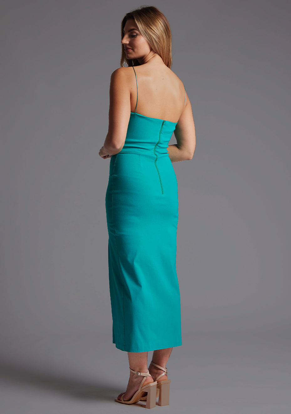 Back quarter image of a model wearing a peacock midaxi dress with a minimalistic strap details, front skirt split and centre back zip. The design featured is the Vesper Tate Peacock Midaxi Dress