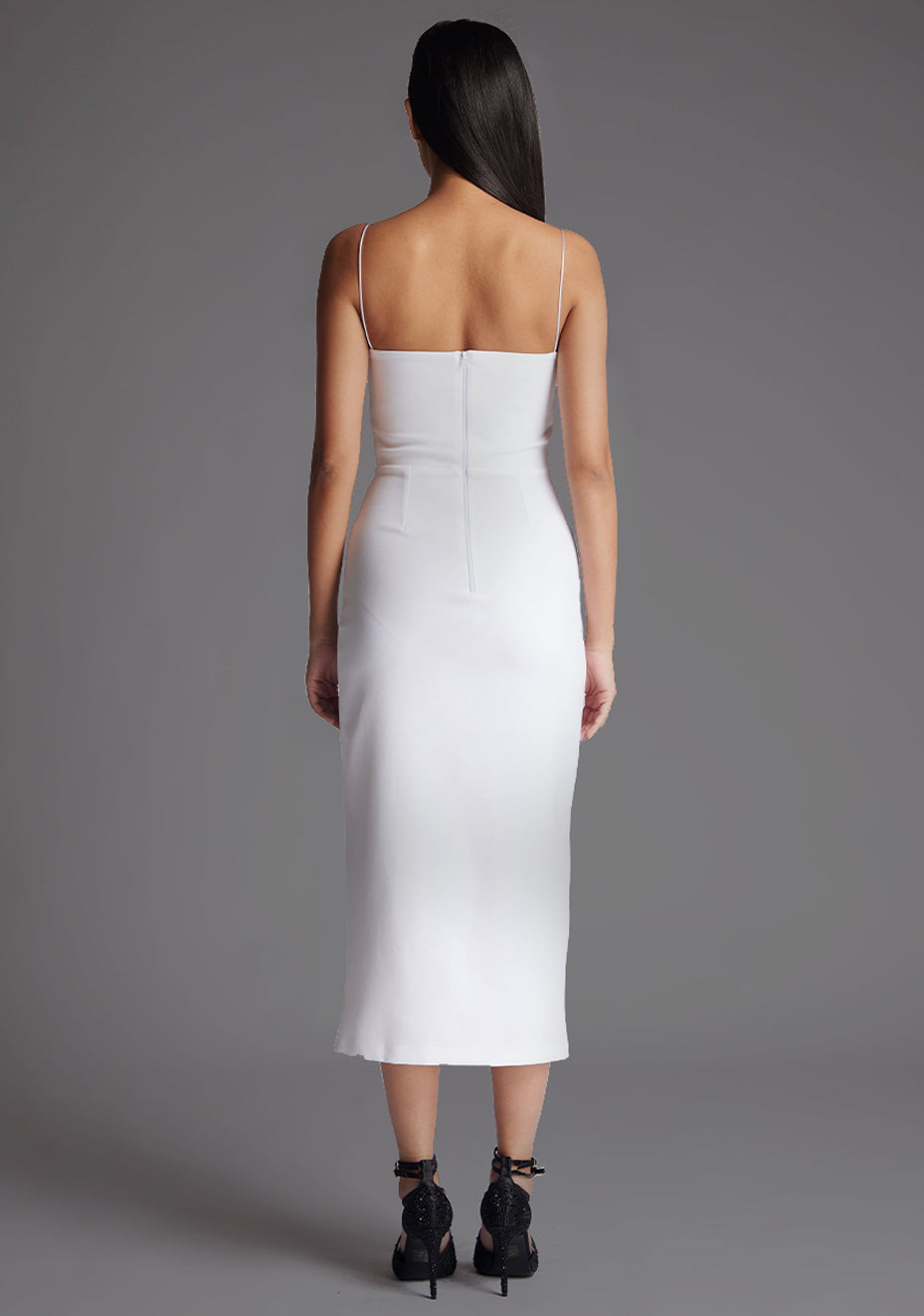 Back image of a model wearing a white midaxi dress with a minimalistic strap details, front skirt split and centre back zip. The design featured is the Vesper Tate White Midaxi Dress