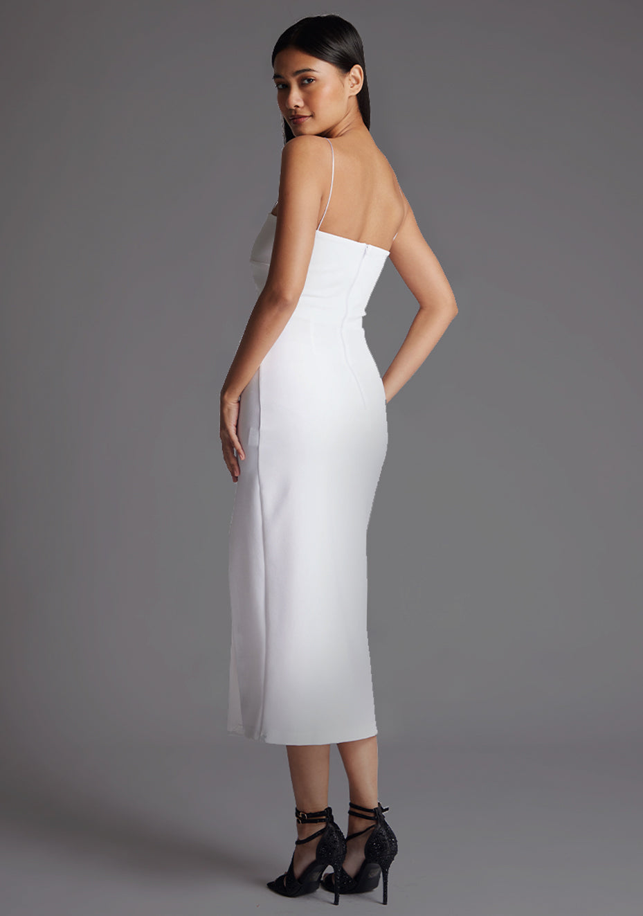 Back quarter image of a model wearing a white midaxi dress with a minimalistic strap details, front skirt split and centre back zip. The design featured is the Vesper Tate White Midaxi Dress