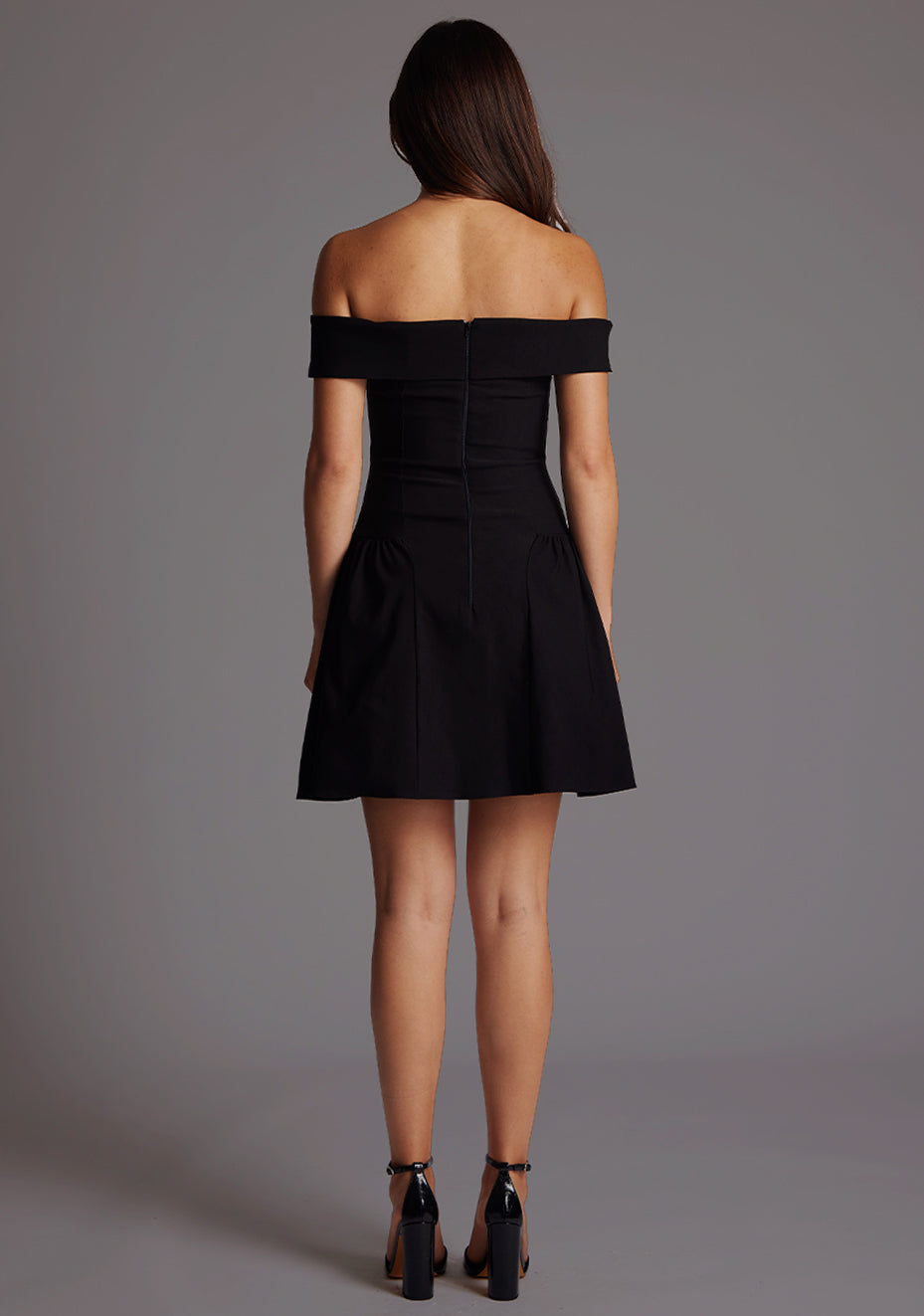Back image of a model wearing a black mini dress with a straight neckline, with an off-shoulder design, bold bardot brand across the arm and neckline and fitted bodice with A-line skirt and centre back invisible zip. The design featured is the Vesper Una Black Mini Dress.