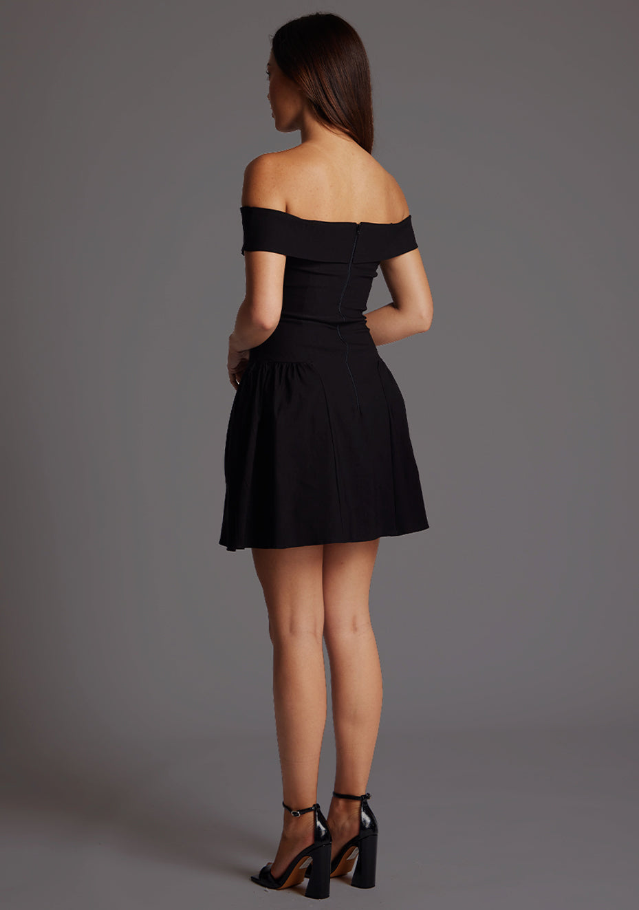 Back quarter image of a model wearing a black mini dress with a straight neckline, with an off-shoulder design, bold bardot brand across the arm and neckline and fitted bodice with A-line skirt and centre back invisible zip. The design featured is the Vesper Una Black Mini Dress