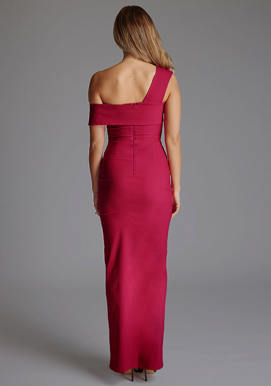 Back image of blonde model wearing a Raspberry Maxi Dress with a asymmetrical silhouette, front skirt split and a bodycon fit. The design features is the Vesper Winslow Raspberry Maxi Dress
