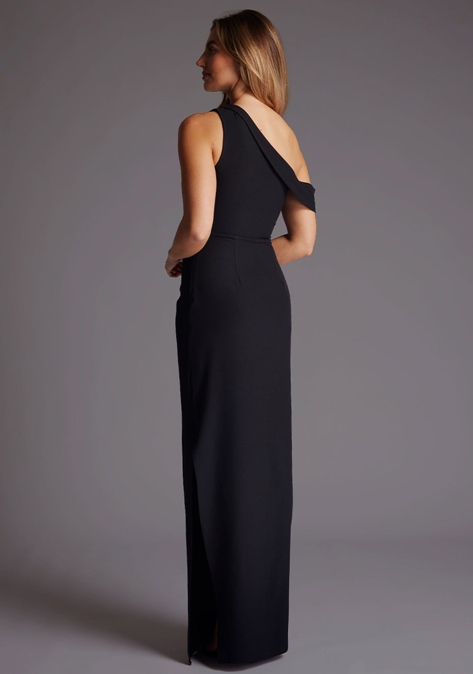 Back quarter image of the model wearing a black maxi dress with asymmetric straps and a side skirt split. The design featured is the Vesper Yvette Black Maxi Evening Dress.