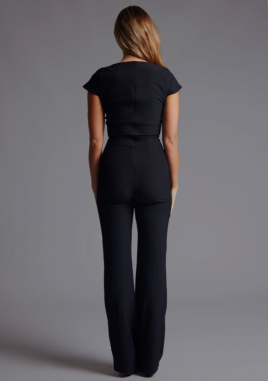 Back image of a model wearing a black jumpsuit with a square neckline and flattering v-wire design, this jumpsuit will have you feeling confident and stylish. The design features is the Vesper Zuri Black Wide Leg Jumpsuit