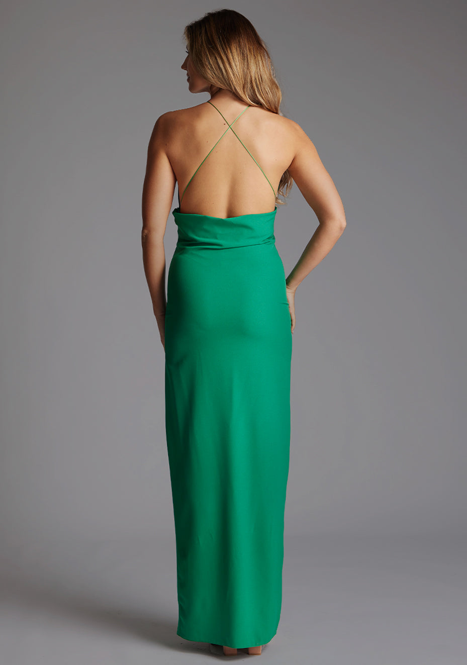 Back image of a model wearing an emerald green maxi dress with a flattering halter neck and a daring front skirt split. The design featured is the Vesper Allegra Emerald Green Maxi Dress