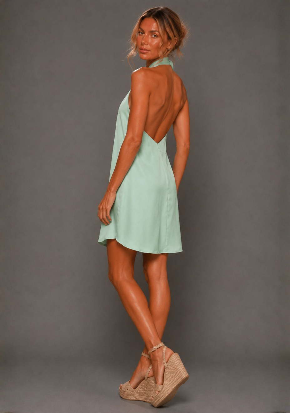 Side back image of a model wearing the VSPR Ayesha Twist Neck Sage Mini Dress in Sage Green, capturing the low V-back detail and graceful movement of the silhouette, standing against a grey studio backdrop