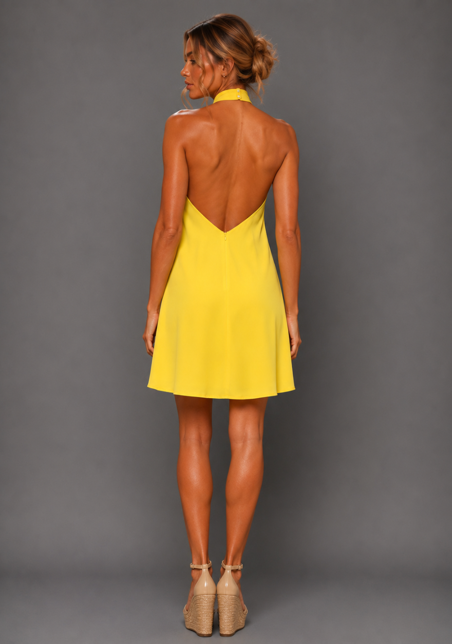 Back image of a model wearing the VSPR VSPR Ayesha Twist Neck Yellow Mini Dress, featuring the contemporary low V-back and graceful loose-fitting silhouette against a grey studio backdrop.