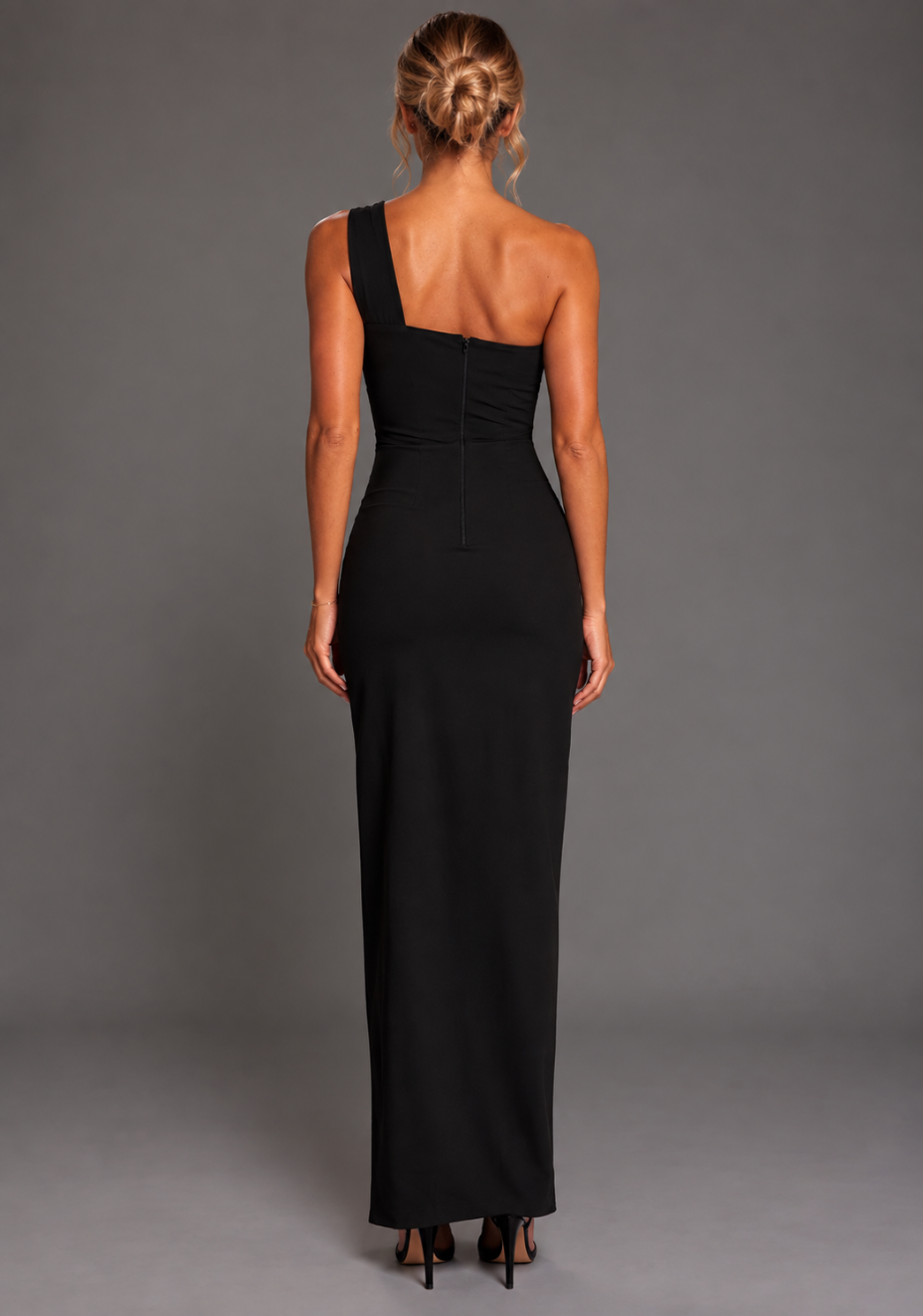 Back image of a model wearing the VSPR Ellis Black Maxi Dress highlighting its sleek one-shoulder styling and graceful maxi-length skirt against a grey studio background.