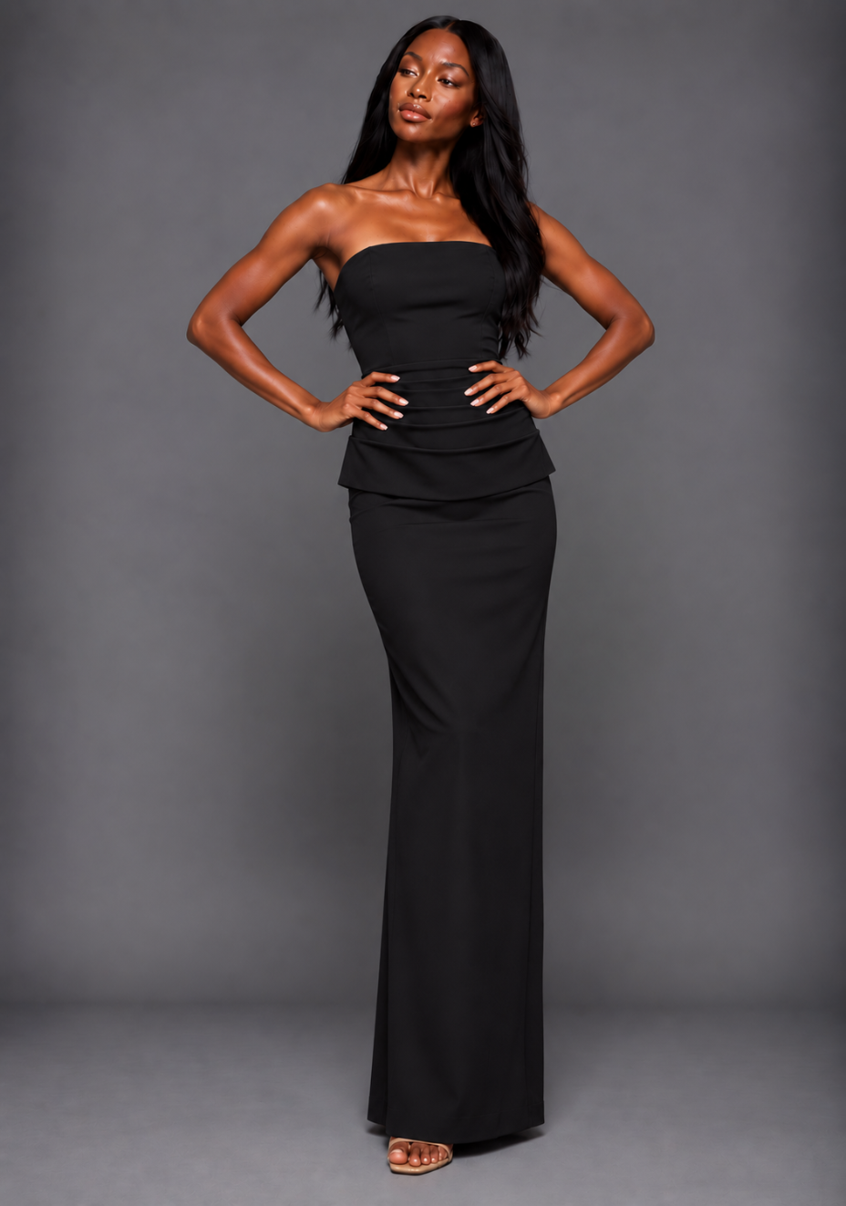 Front image of a model wearing the VSPR Isabel Bandeau Ruched Waist Detail Black Maxi Dress, featuring a strapless neckline, ruched waist and floor-length skirt against a grey studio backdrop.