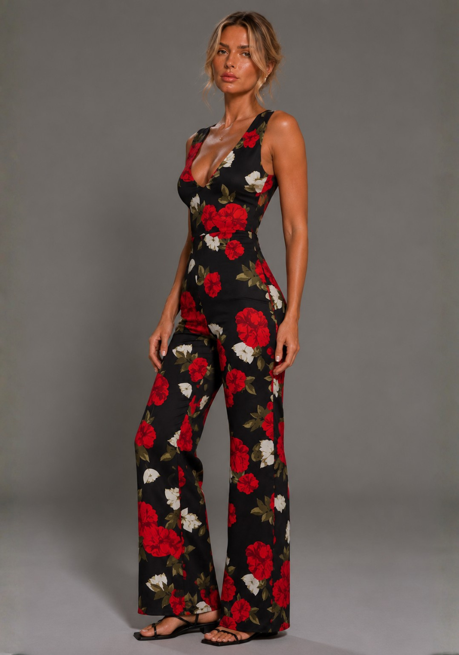Side front image of a model wearing the VSPR Noelle V Neckline Black Floral Wide Leg Jumpsuit highlighting the plunge neckline, contoured waist, and flowing wide leg trousers against a grey studio backdrop.