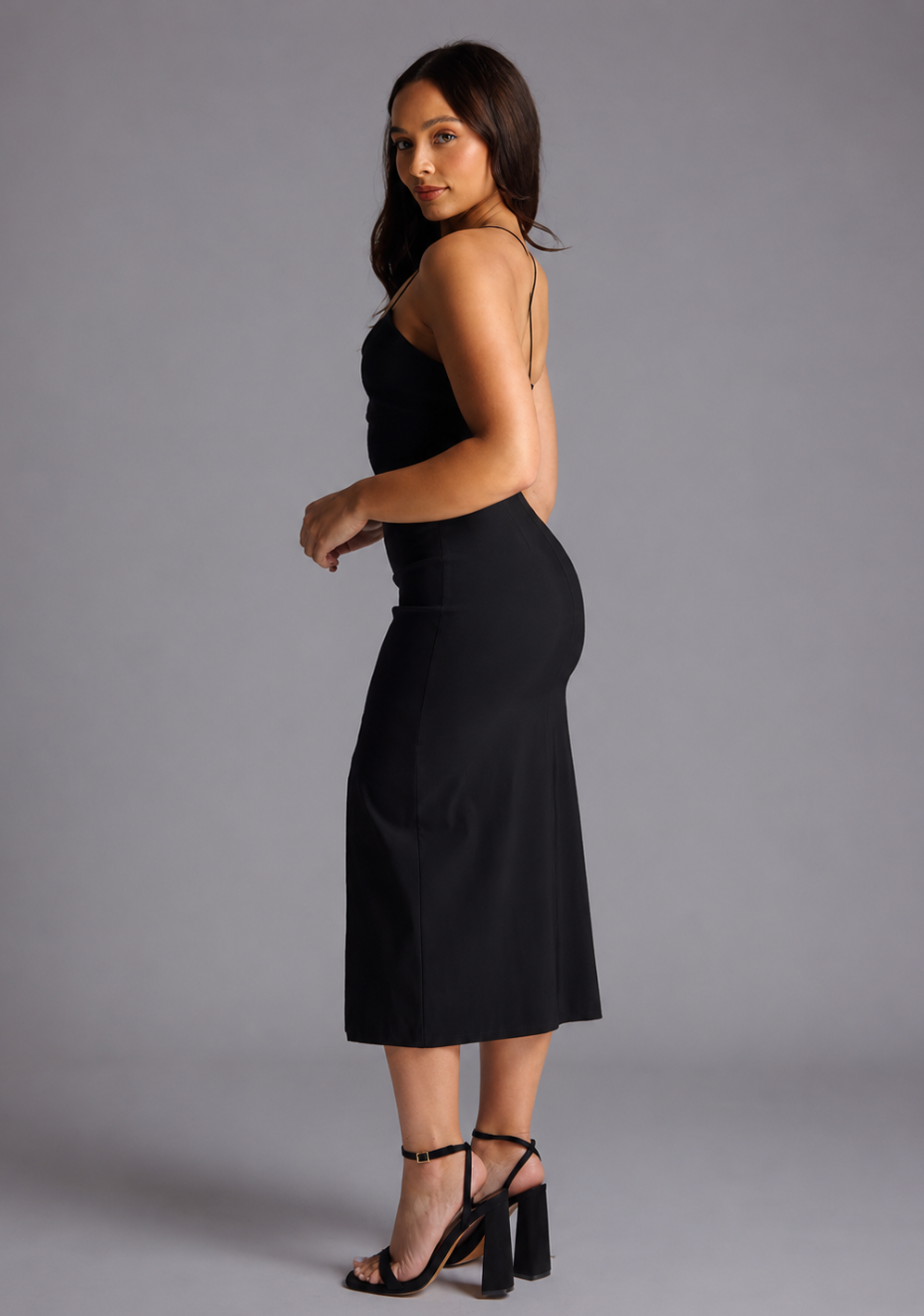 Woman wearing the Vesper Petite beryl black midaxi dress with a thigh slit on a grey background, side back view