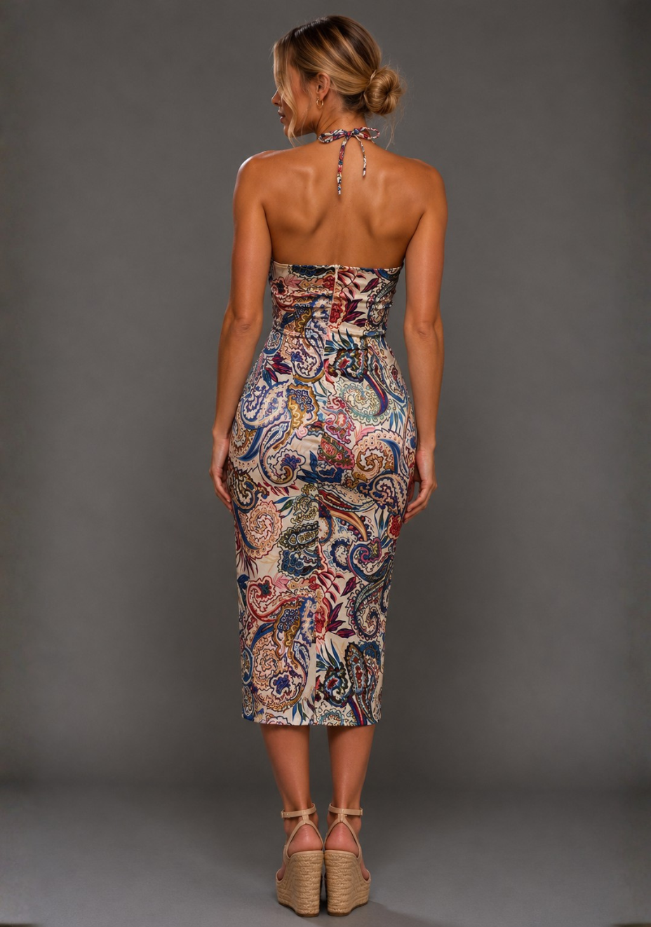 Back image of a model wearing the VSPR Pandora Paisley Print Midi Dress in Paisley Print with a halterneck tie fastening and sculpted silhouette, standing against a grey studio backdrop