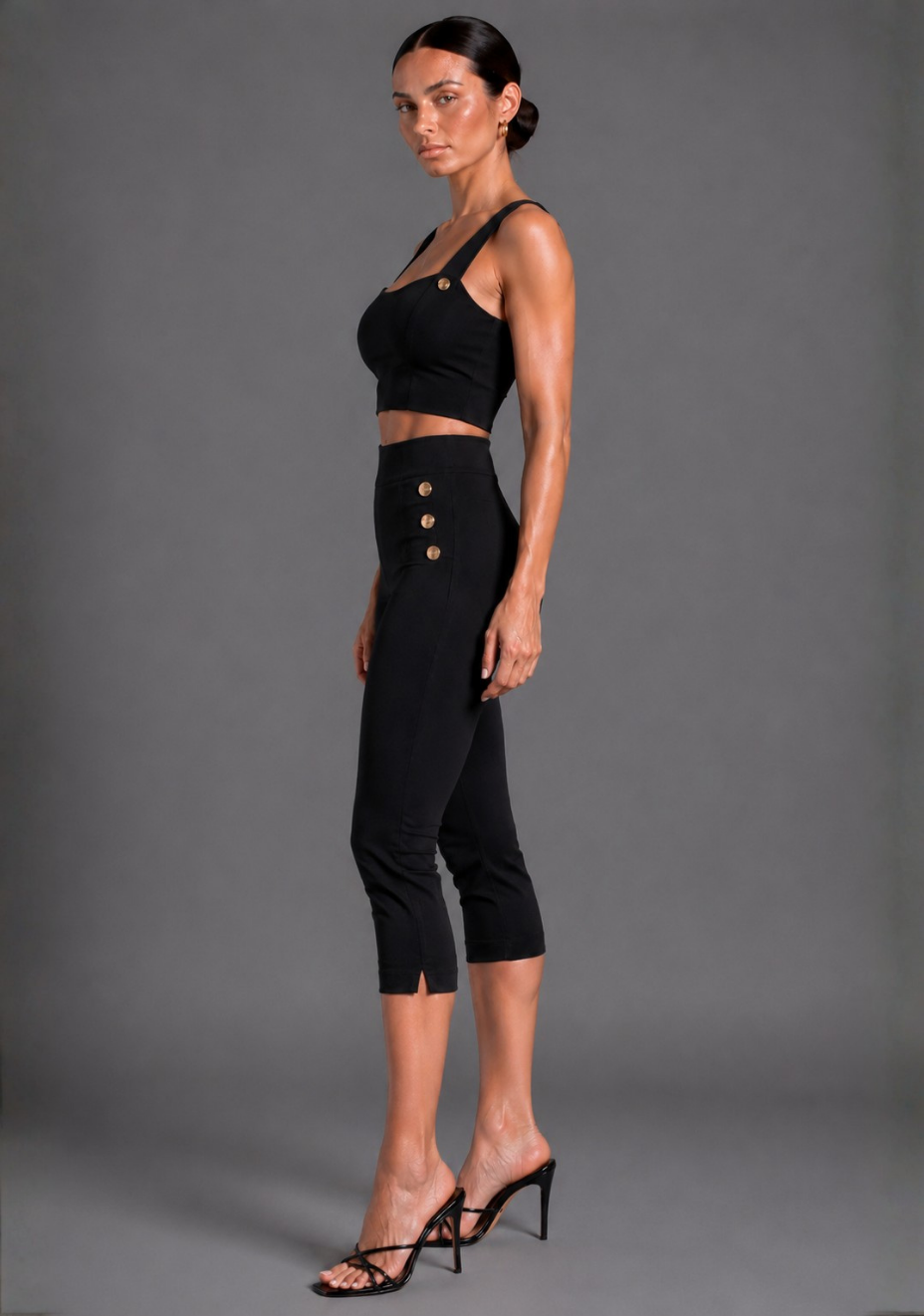 A woman modeling a black two-piece outfit consisting of a sleeveless crop top with gold button details and matching high-waisted capri pants with gold buttons down the side, paired with black strappy heels, against a solid gray studio background.