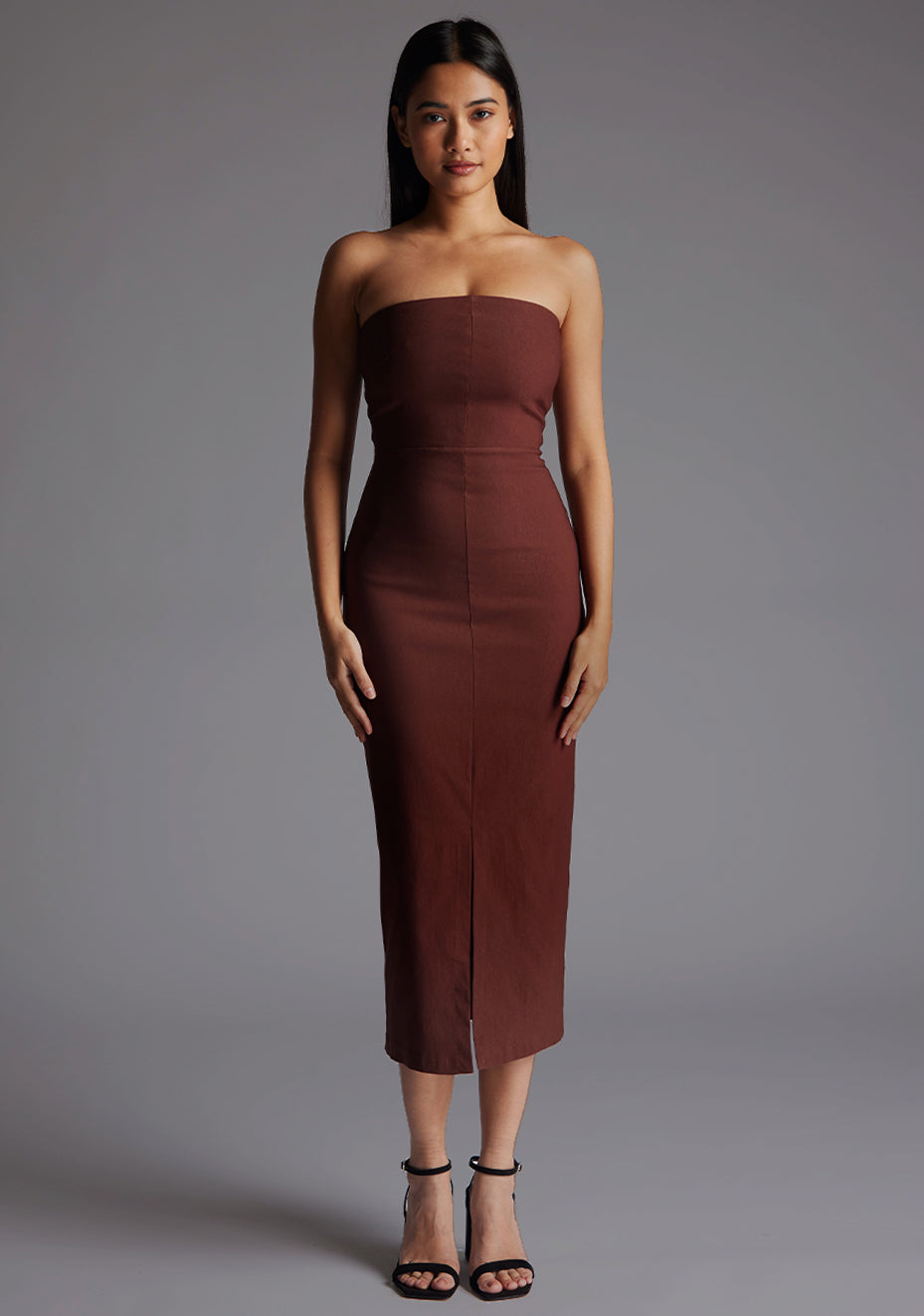Front image of a model wearing the Vesper Andorra Strapless Midaxi Dress in Chocolate, featuring a sleek strapless neckline, slim-fit silhouette, and elegant front skirt split, posed against a grey studio backdrop.