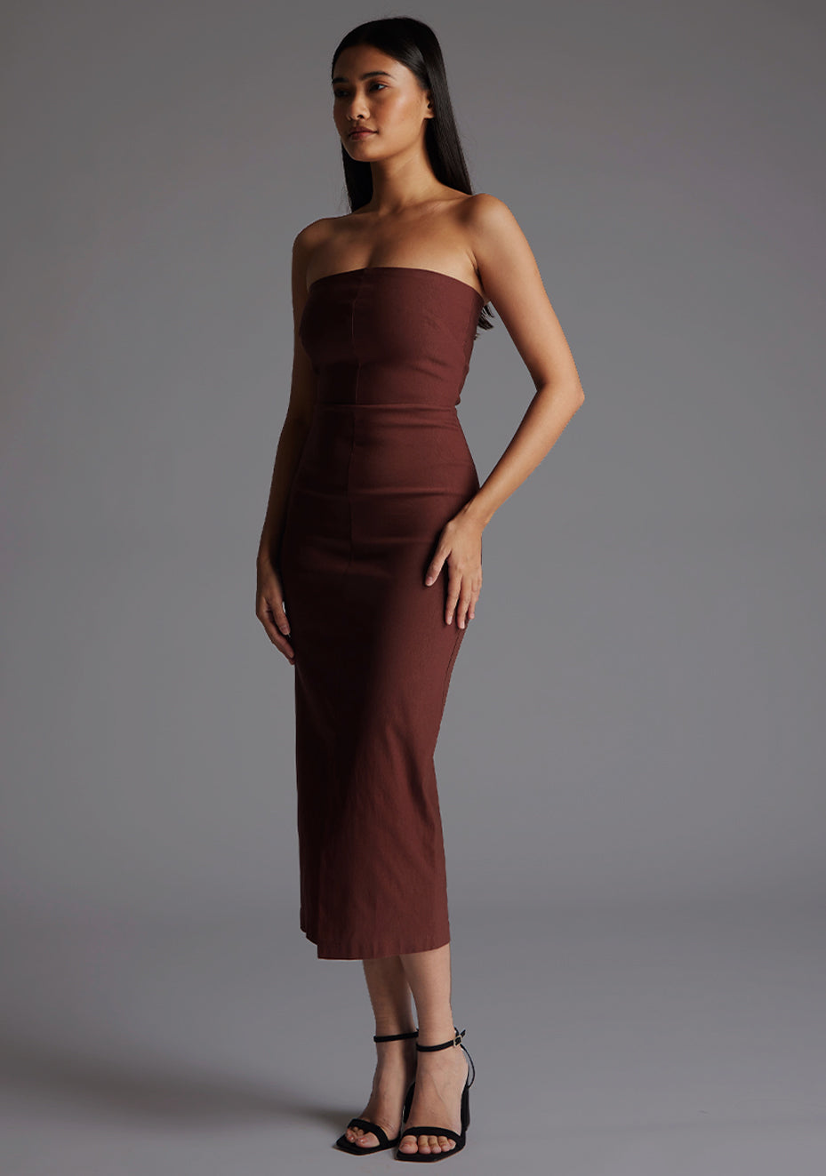 Side front of a model wearing the Vesper Andorra Strapless Midaxi Dress in Chocolate, highlighting the body-contouring fit, smooth strapless bodice, and refined midaxi length in a minimalist studio setting.