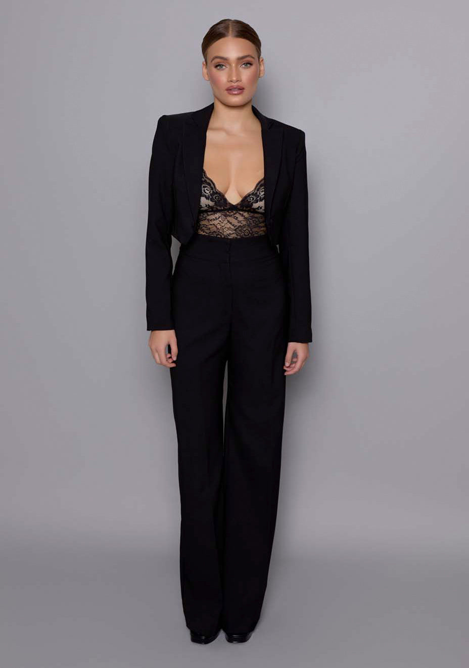 Model wearing the VSPR Aria Cropped Tailored Jacket in black, featuring a structured silhouette with sharp lapels and tailored shoulders, styled with matching high-waist trousers against a grey studio backdrop.