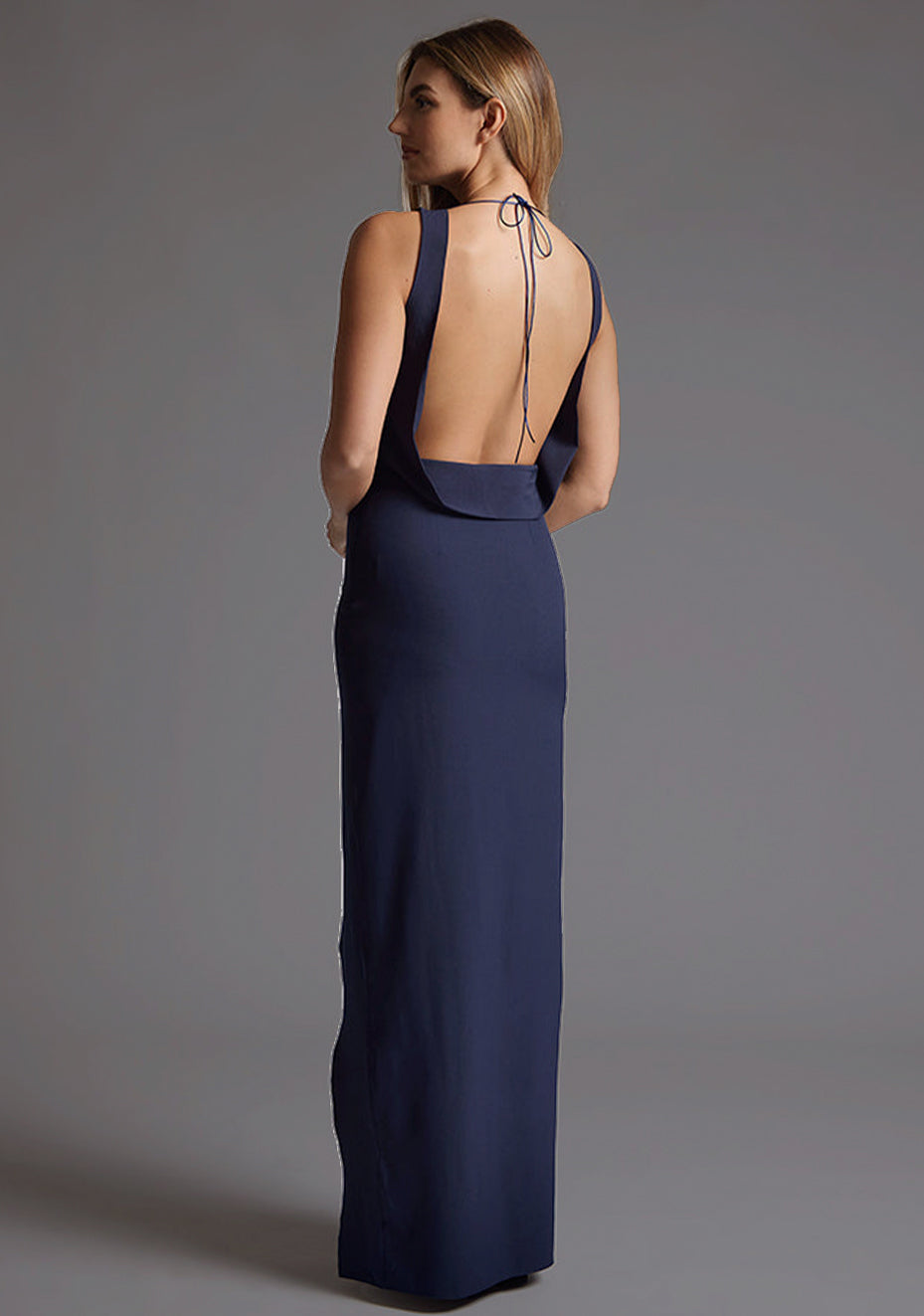 Side back image of a model wearing the Vesper Brodi Backless High Neck Maxi Dress in Navy, revealing the open back and smooth tailored fit.