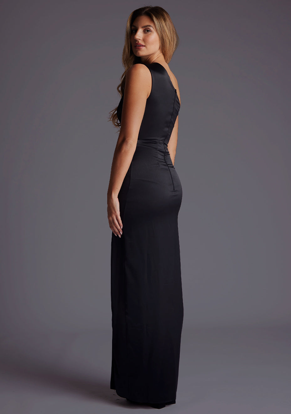 Side back image of a blonde model wearing a black maxi dress with an alluring asymmetric one-shoulder design, subtle side cut-out and flattering wrap design skirt. The design featured is the Vesper Candace One-Shoulder Satin Maxi Dress in Black