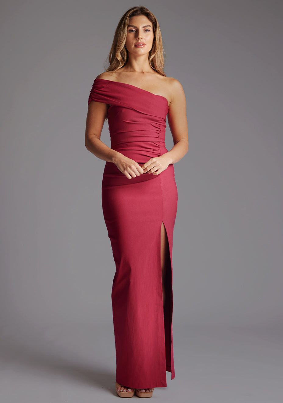 Front image of a model wearing the Vesper Carmel Off-the-Shoulder Maxi Dress in Raspberry, featuring a one-sleeve neckline, ruched bodice, and dramatic thigh-high slit, standing against a grey studio backdrop.