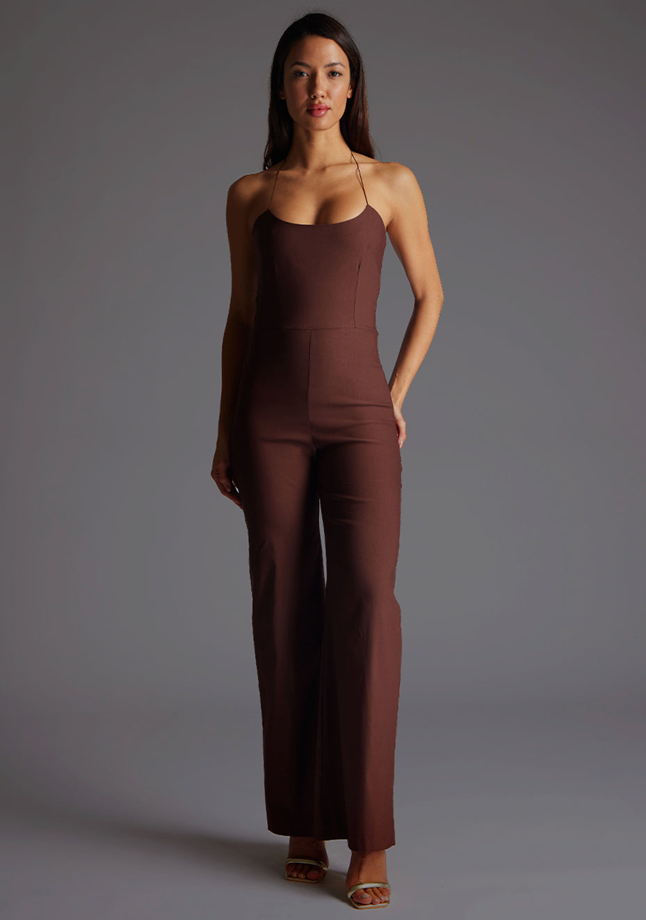 Front image of a model wearing the Vesper Cassidy Halterneck Wide Leg Jumpsuit in Chocolate, featuring a sleek halterneck design and wide-leg silhouette, standing against a grey studio backdrop.
