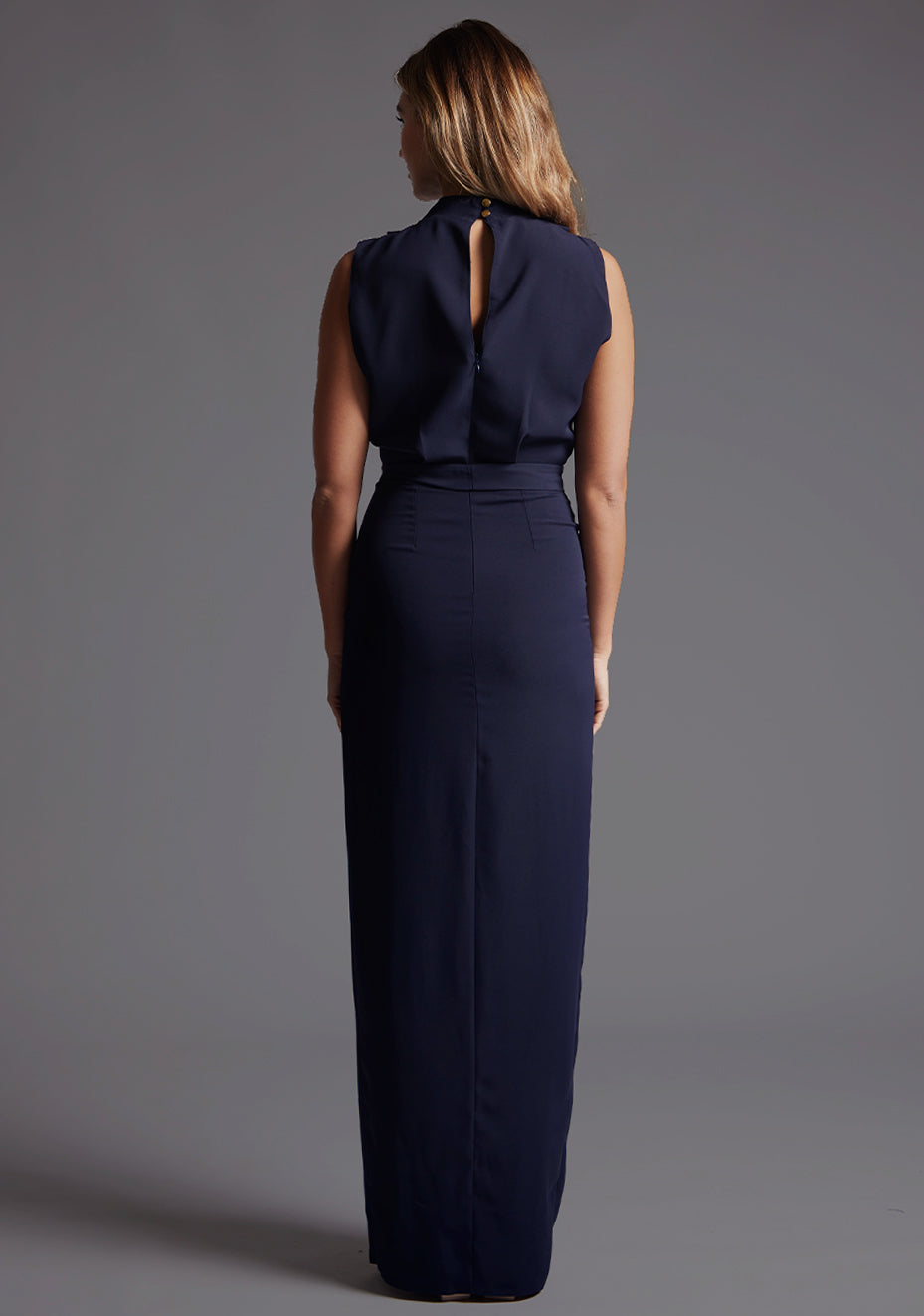 Back image of a model wearing the Vesper Chrysta High Neck Maxi Dress in Navy, featuring a concealed zip closure and sleek, uninterrupted silhouette against a grey studio backdrop.