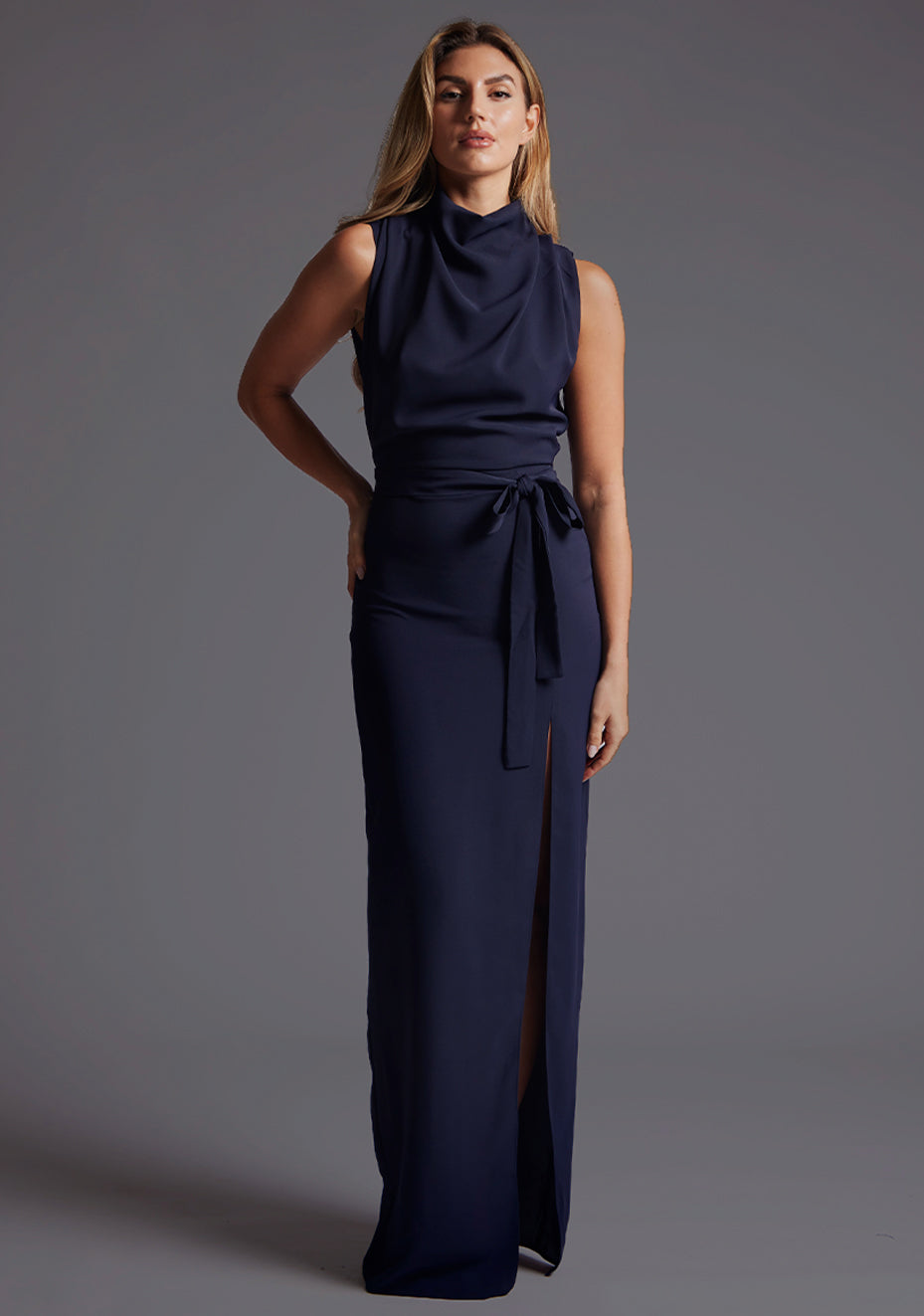 Front image of a model wearing the Vesper Chrysta High Neck Maxi Dress in Navy with a slim-fit silhouette, high neckline, and subtle front skirt split, standing against a grey studio backdrop.
