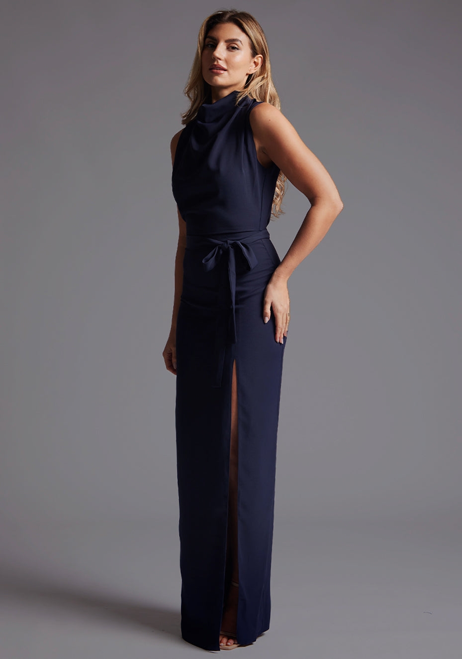 Side front image of a model wearing the Vesper Chrysta High Neck Maxi Dress in Navy, revealing the slim profile and gentle movement created by the front skirt split.