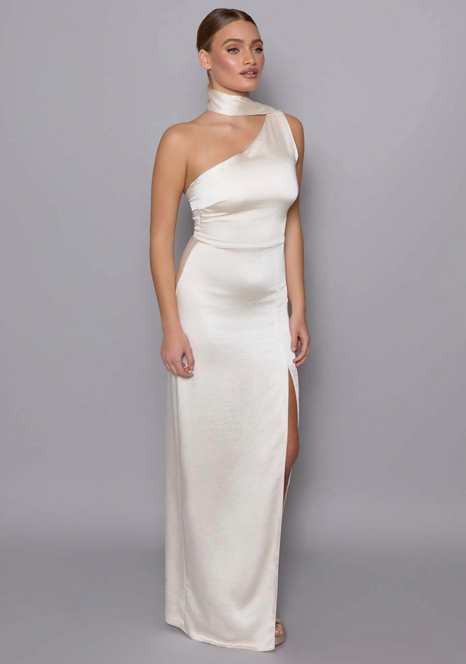 Side-front view of the VSPR Cordula cream maxi dress emphasizing the one-shoulder neckline, body-skimming fit, and flowing satin scarf detail that cascades gracefully.