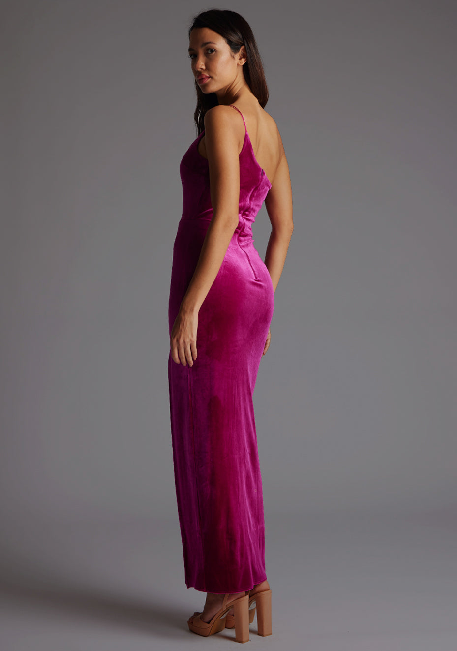 Side back model wearing a magenta maxi dress with a flattering asymmetric cut-out neckline, finished with a one shoulder strappy detail and front skirt split. The design featured is the Vesper Daniella One Shoulder Velvet Maxi Dress Magenta.