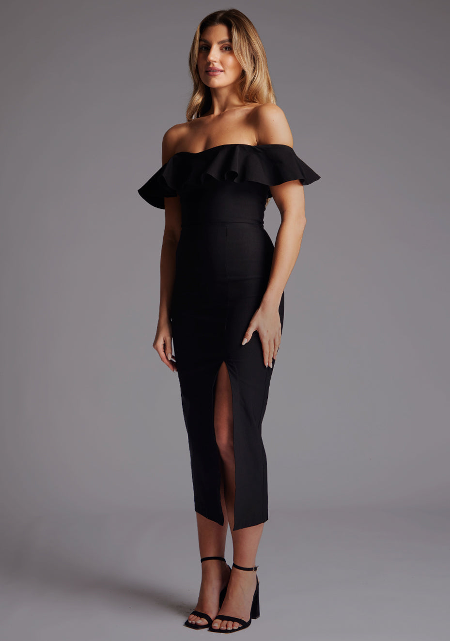 Side front image of a model wearing a black midaxi dress with a bardot neckline with a ruffle design, front skirt split and invisible centre back zip. The design featured is the Vesper Deirdre Bardot Off-Shoulder Frill Midaxi Dress in Black.