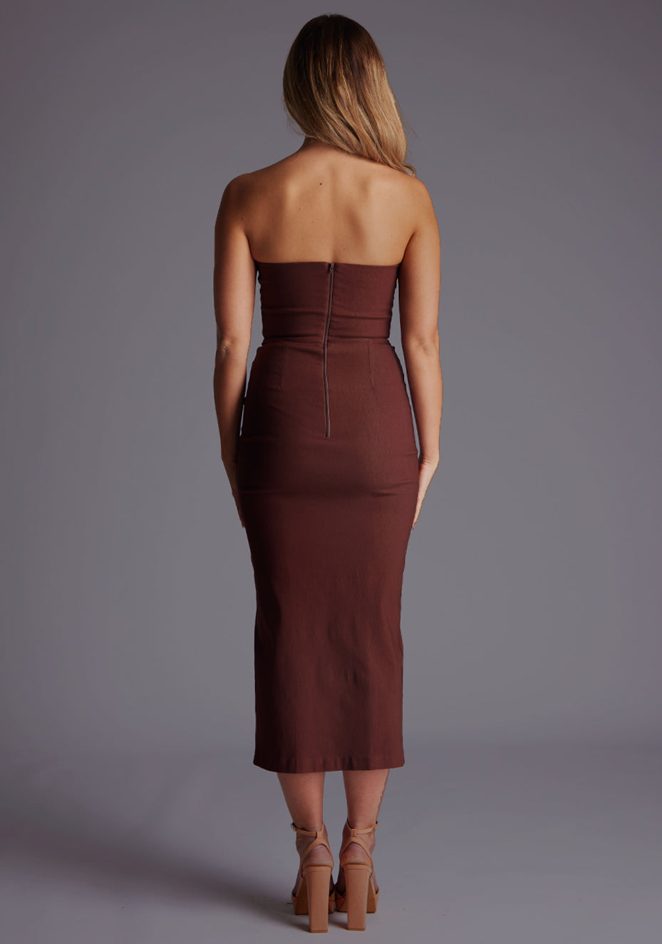 Back  image of a blonde model wearing a chocolate midaxi dress with a strapless gown, front cut-out that adds a touch of allure, while the front skirt split adds a modern flair. The design featured is the Vesper Delaney Strapless V-Front Midaxi Dress Chocolate..