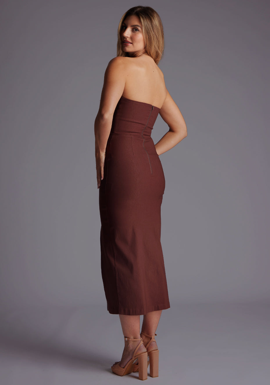 Side back image of a blonde model wearing a chocolate midaxi dress with a strapless gown, front cut-out that adds a touch of allure, while the front skirt split adds a modern flair. The design featured is the Vesper Delaney Strapless V-Front Midaxi Dress Chocolate.