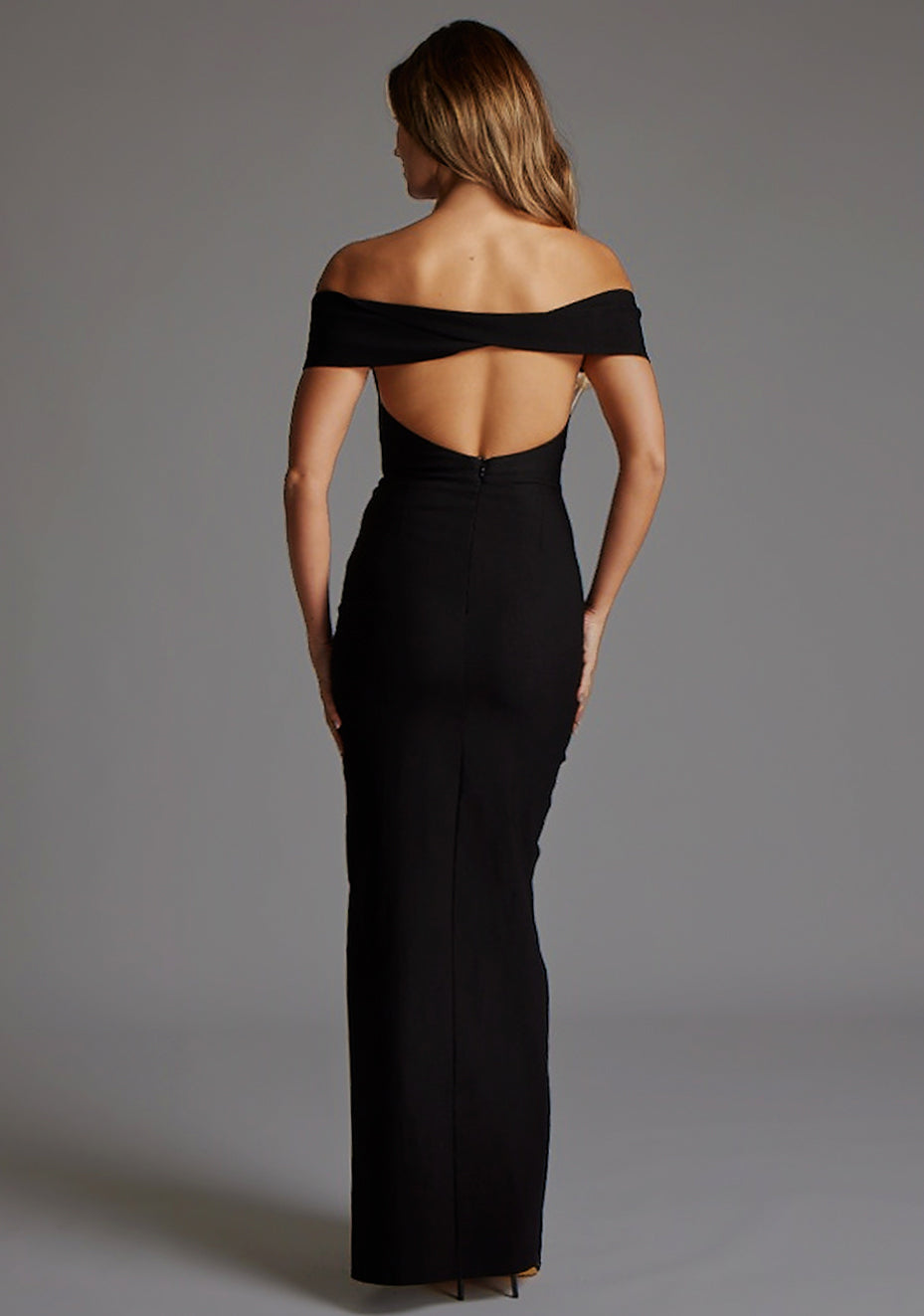Back image of a model wearing the Vesper Delta Bardot Low Back Maxi Dress in Black, highlighting the dramatic low back detail and clean, elongated silhouette.
