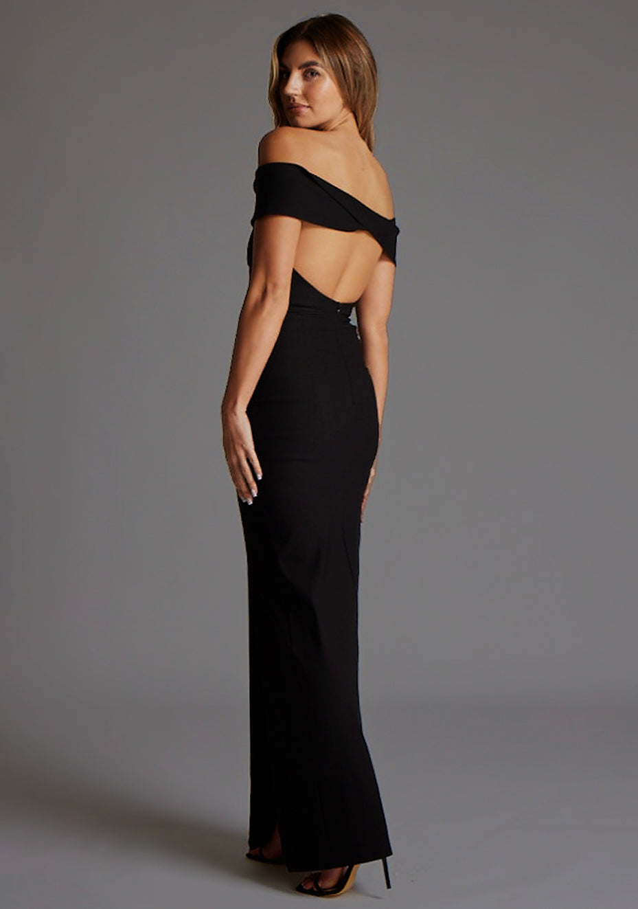 Side back image of a model wearing the Vesper Delta Bardot Low Back Maxi Dress in Black, revealing the low back detail and streamlined bodycon shape.
