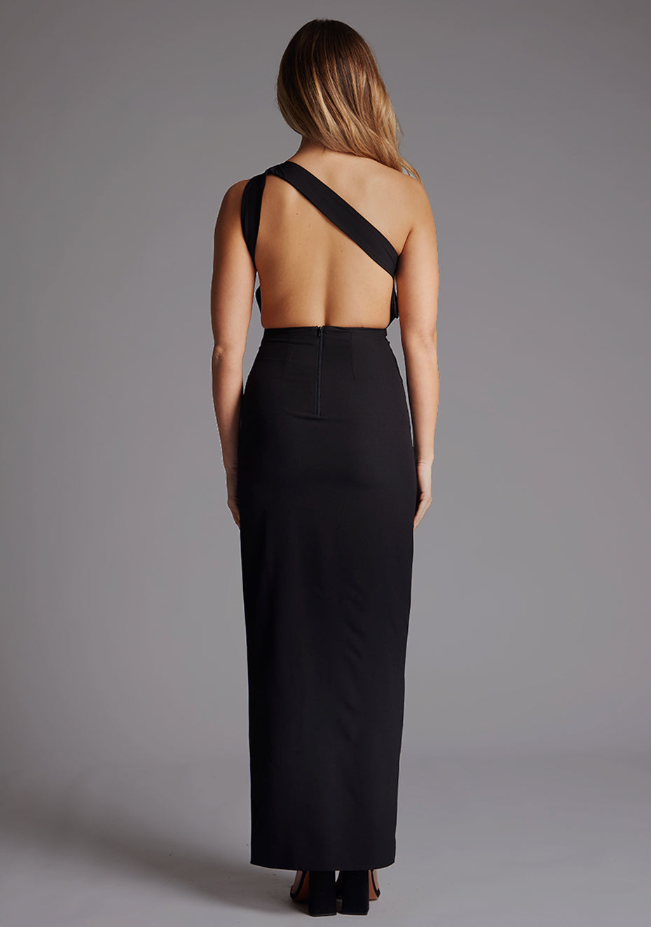 Back image of a model wearing the VSPR Deva One Shoulder Open Back Black Maxi Dress, showcasing the striking open-back design and flowing maxi length against a grey studio backdrop.