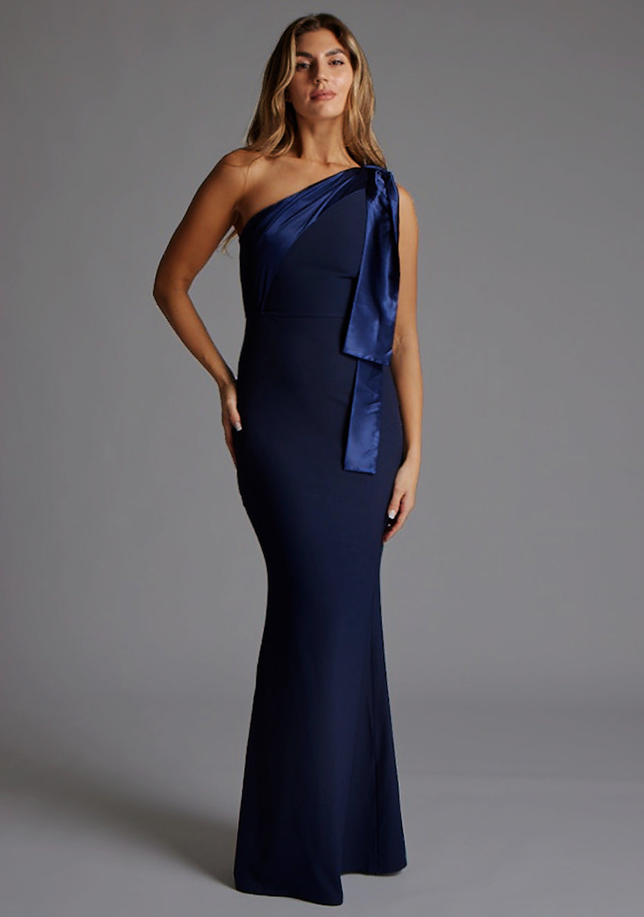 Front image of a blonde model wearing a Navy Maxi Dress with an asymmetric neckline and one-shoulder. The design features the Vesper Deven One Shoulder Fishtail Maxi Dress in Navy.